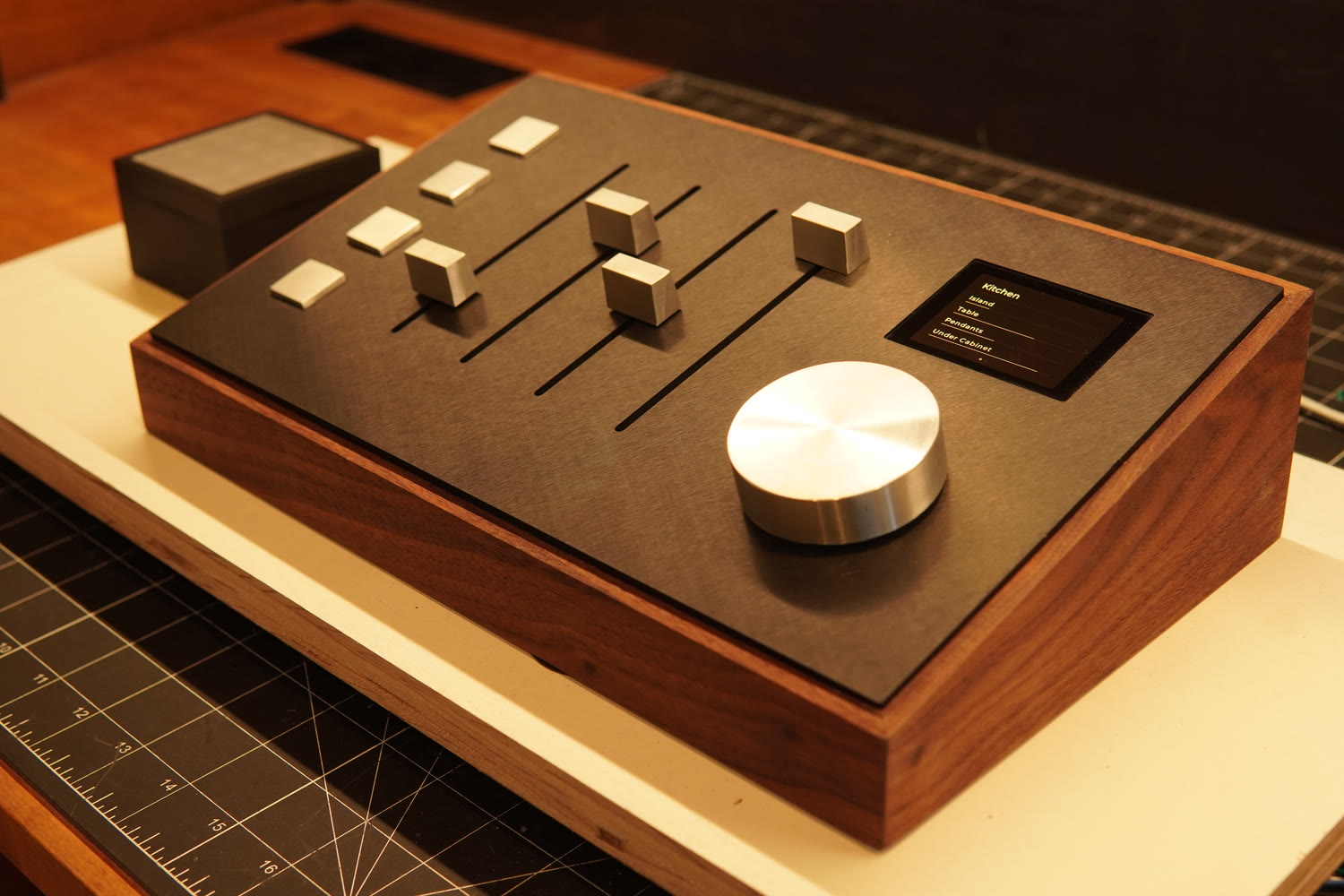 A walnut-bodied lighting console with a dark faceplate, four aluminum fader caps, a small display and a large spun-aluminum knob, on a workshop bench.