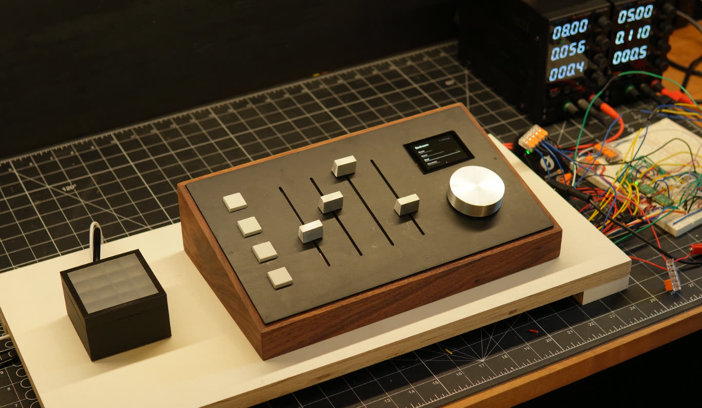 A walnut-bodied lighting console with a dark faceplate, four aluminum fader caps, a small display and a large spun-aluminum knob, on a workshop bench.