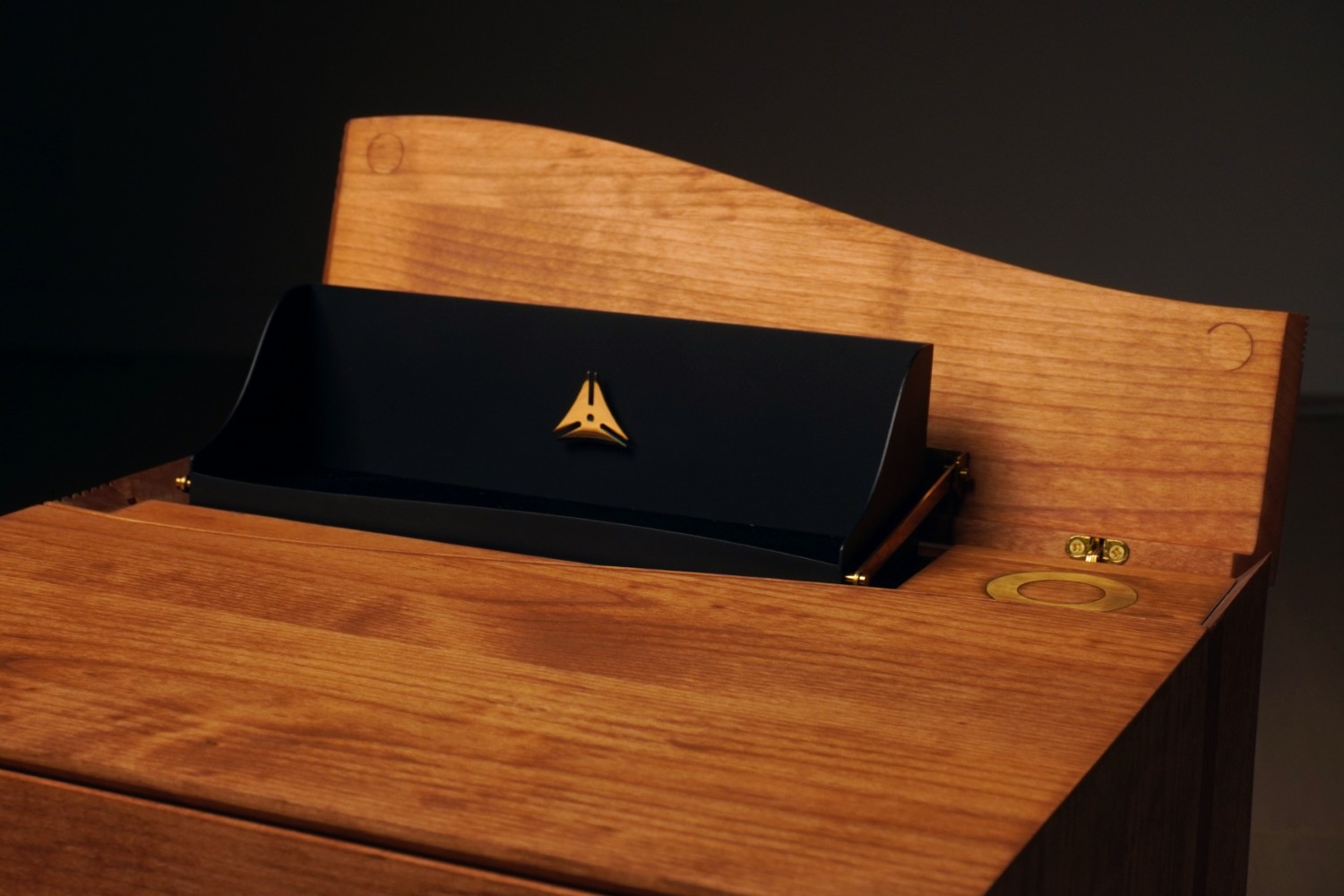 The top of a wooden nightstand, opened to show a black steel tray with a brass Sacrebleu logo, raised on a brass hinge.